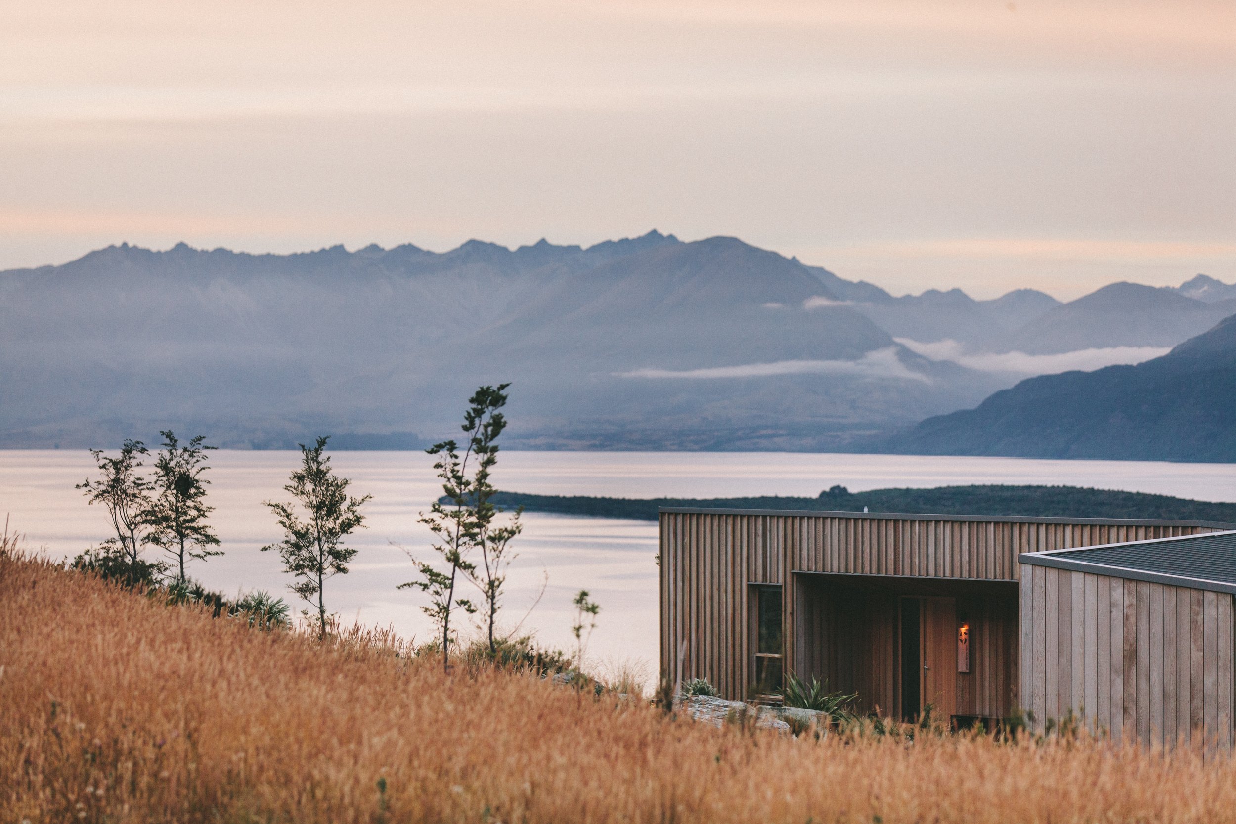 Modern wooden house with large windows and a sloped roof, surrounded by tall grasses and a few trees, with a large lake and distant mountains in the background at sunset.